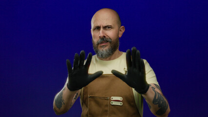 Man with beard and tattoos wearing gloves and overalls looks concerned against an isolated blue background