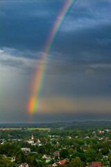 A vivid rainbow brightens the stormy sky above a green town landscape with a church tower in the foreground