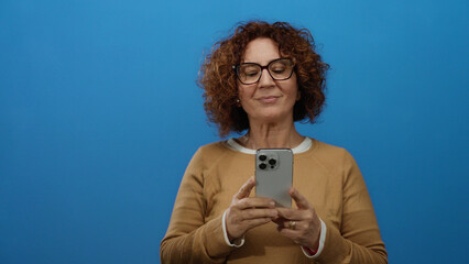 Middle aged woman with curly hair in glasses using smartphone against a blue background wall, wearing brown sweater, showing thoughtful expression and hispanic ethnicity.