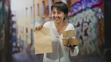 Woman holding paper bag and stacked takeout containers, hand visible in a street alley with graffiti; community sharing joy.