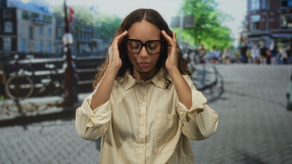 Fototapeta premium Woman with glasses touching head and hand on chest on urban street by canal and bridge; fatigue contemplation.