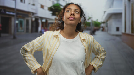 Fototapeta premium Woman touching chin with finger on a city street in a casual striped shirt, glancing upward; thoughtful curiosity.