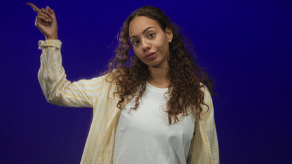 Woman with raised arm showing thumbs down, wearing yellow striped shirt and white tee in a blue studio; disapproval.