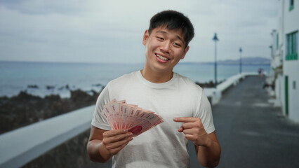 Young man smiling and holding taiwan dollars on a seaside promenade with an ocean view.