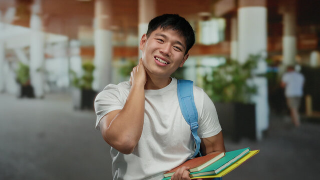 Young man outdoors holding books with a confused expression, wearing a blue backpack on a city street with people in the background and greenery around.