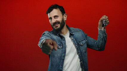 Hispanic man in denim jacket excitedly holding car key with vivid red background pointing at camera...