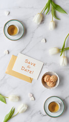 Save the Date card on a marble tabletop arranged with white tulips, two teacups and small treats with ample copyspace and available space for text, offering a clean layout for invitations and seasonal