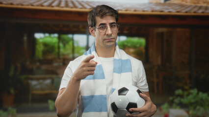 Man wearing light blue and white scarf and glasses, holding a soccer ball in left hand and pointing...