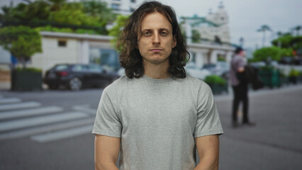 Fototapeta premium Man with long hair standing with arms relaxed on street near crosswalk and parked car; introspection solitude.