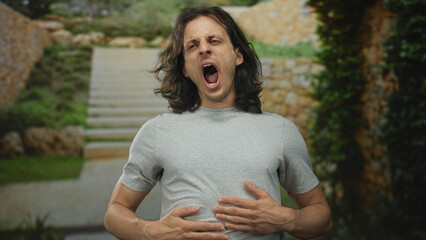 Young hispanic man with long hair winking and puckering lips in front of stone building stairs outdoors wearing gray t shirt; playful mischief.