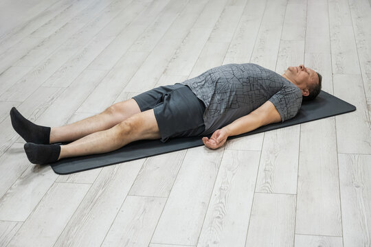 Adult man lying in shavasana pose on yoga mat during relaxation practice, improving mindfulness, breathing, and body recovery in calm studio.