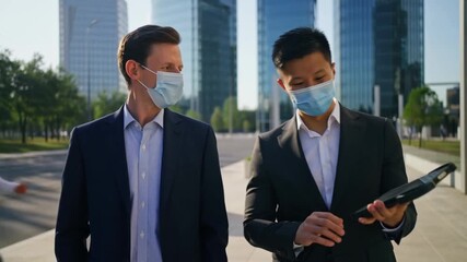 Two businessmen, wearing face masks, walking outdoors and discussing their work in front of modern skyscrapers. The atmosphere is professional and the scene suggests a business discussion. Stock Video - Powered by Adobe
