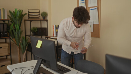 Young man with beard reviewing documents in modern office surrounded by charts, computer, and plants, showcasing a focused work environment.