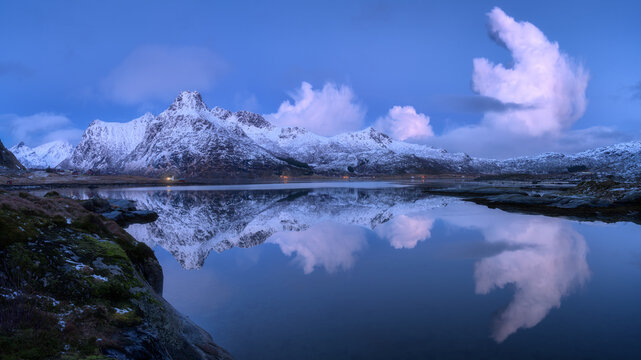 Snow-covered mountains reflected in a calm fjord near a small village on the Lofoten Islands in Norway. Winter landscape with rorbu houses, shoreline and still water in cold evening light at dusk - Powered by Adobe