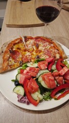 A plate with pepperoni pizza and a fresh vegetable salad is served with a glass of red wine on a wooden table. The scene conveys cozy dining, comfort food and a relaxed evening atmosphere
