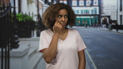 Fototapeta premium Woman in pale pink shirt points finger to lower eyelid in a city street outside shops; health concern.