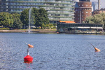 Red and Orange Water Buoys Floating in El&auml;intarhanlahti bay in the Finnish capital Helsinki. Hakaniemi silhouette with a Fountain and a restaurant in Background.