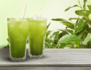 Matcha drink with ice in glasses on wooden table against green leaves. Space for text