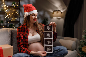 A woman wearing a Santa hat holds ultrasound images, celebrating the upcoming arrival of her baby during the Christmas season