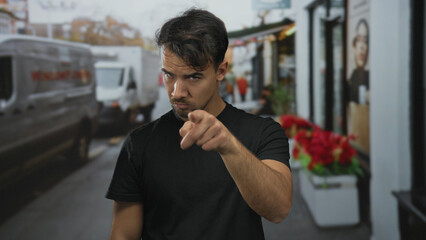 Young hispanic man in black shirt makes a watchful gesture on a bustling city street lined with...
