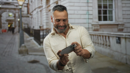 Man holding smartphone with both hands on a street, smiling and gesturing while looking at screen; digital connection amusement.