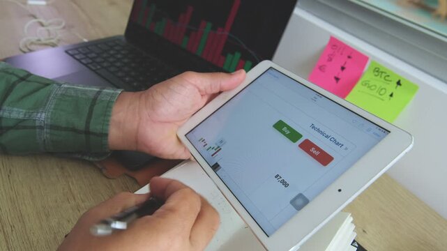 Crypto trader analyzing stock market chart on a digital tablet and taking notes in a notebook
