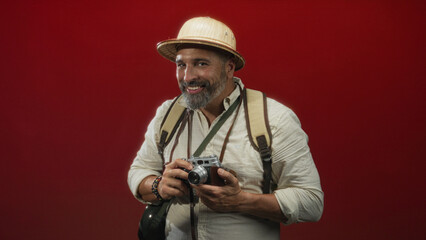Man holding a vintage camera and wearing a pith helmet and backpack in studio with red backdrop; adventure curiosity.