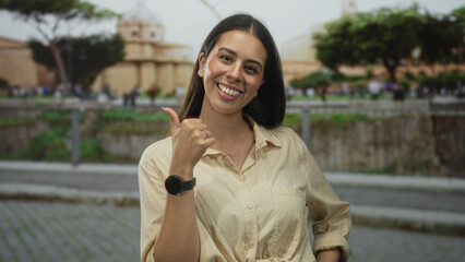 Woman showing thumbs up with raised hand and visible smartwatch in front of a stone building, smiling; confidence approval.