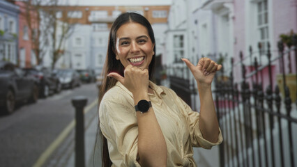 Woman, young brunette, thumbs pointing to self showing smartwatch on wrist on a city street lined with pastel townhouses and parked cars; confidence celebration.