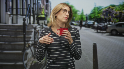 Woman holding red mug with hands and looking aside, glasses on, striped top on a street lined with bicycles and parked cars; quiet contemplation.
