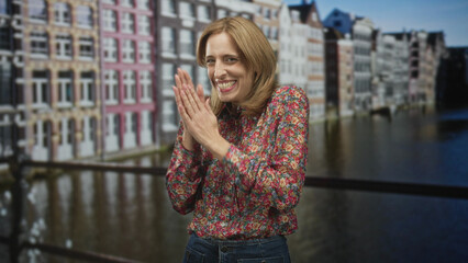 Fototapeta premium Woman clapping hands on a bridge by a canal in front of colorful historic buildings, wearing floral blouse and jeans; joy celebration.