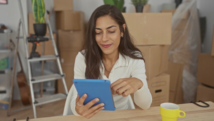 Woman smiling while tapping tablet with hands among moving boxes and ladder in building; home productivity contentment.