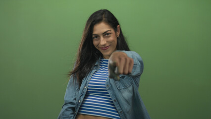 Woman points finger at camera in green studio wearing denim shirt and blue striped crop top; confidence empowerment.