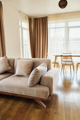 Bright living room with modern sofa and soft cushions on warm wood floor, airy dining area with table and chairs, sheer curtains and warm sunlight creating a cozy contemporary home interior.