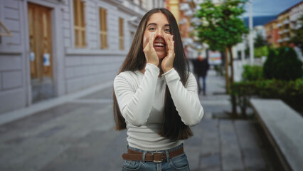 Fototapeta premium Young woman cupping hands around mouth shouting on a pedestrian street by buildings and a bench, wearing white top and jeans; joyful celebration.