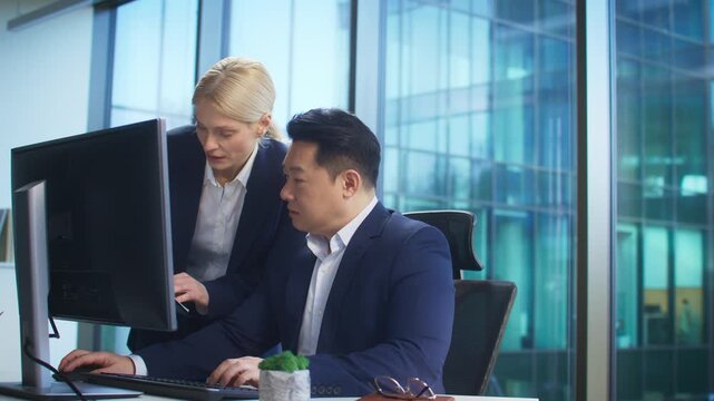Two colleagues in formal suits working together in bright office while comparing information on computer screen and clipboard during focused discussion. Searching for mistake. Teamwork.