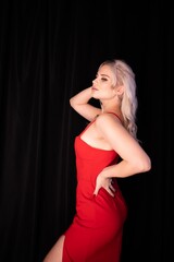 Fototapeta premium Elegant blonde woman in red evening dress posing in dark studio with black backdrop, glamour fashion portrait with dramatic lighting, stylish model in high heels showcasing luxury feminine beauty 