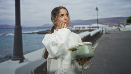 Woman chef holding green pot, hand pointing forward on a seaside street with lampposts and mountains visible; determination decision.