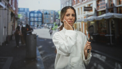 Obraz premium Woman chef holding spatula with hand on cheek showing toothache on street; concerned fatigue stress worry.