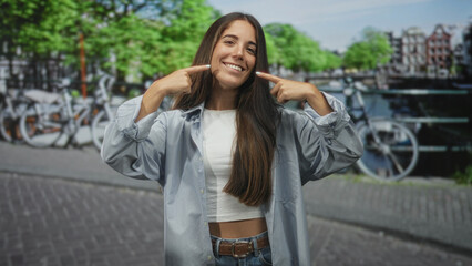 Woman points fingers to her smiling mouth on a street by a canal with bicycles and cobblestones; travel joy memories.