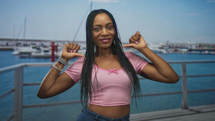 Woman points thumbs to chest showing midriff and smiles wearing pink crop top and bracelets on a...