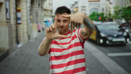 Young man with tattoos making framing gesture on city street wearing red striped shirt outdoors,...