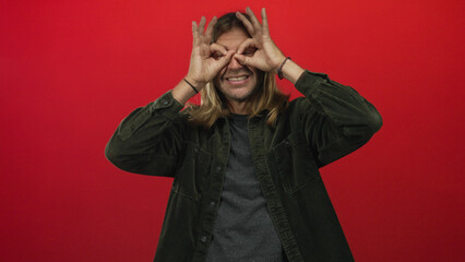 Man wearing corduroy jacket smiles while making hand binoculars gesture in a solid red studio;...