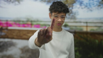 Fototapeta premium Young hispanic man in white sweater and round eyeglasses pointing finger at camera on busy street; skepticism.