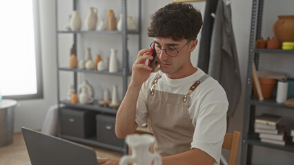 Man holds smartphone to ear in studio among pottery shelves and wears apron as artisan; quiet concentration.