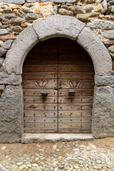 An ancient and robust dark wooden double door, reinforced with metal studs and equipped with stylized heart-shaped carved handles. 