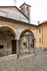 A quaint, rustic-style portico with flaking plaster and stone columns leads to the dark wooden entrance of the Chiesa di San Rocco dell'Ollera in Biella.