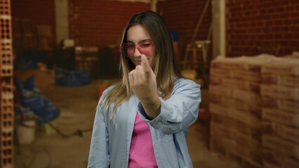 Fototapeta premium Young woman with blonde hair making a beckoning gesture with finger, wearing pink sunglasses at indoor construction site with bricks and tools.