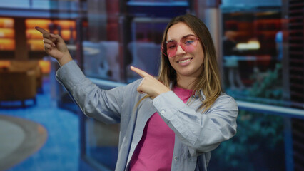 Young woman with blonde hair wearing pink sunglasses and pointing excitedly in a modern hotel...