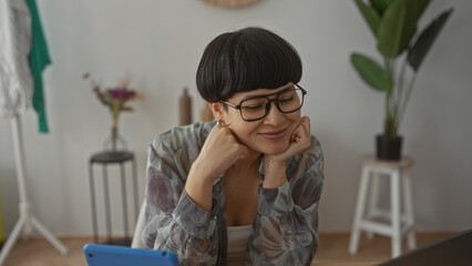 Woman smiling indoors in cozy apartment with plants, smiling while using laptop and tablet, dressed casually, conveying a sense of relaxation and happiness in modern living space.